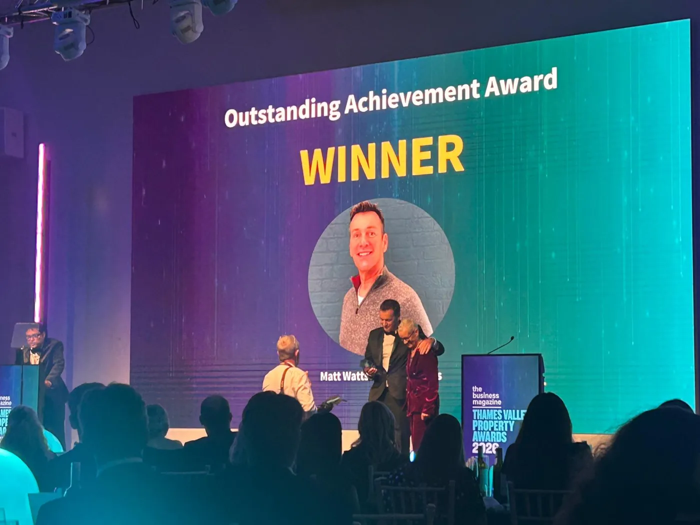 A large screen reading Outstanding Achievement Award, Winner, with a photograph of Matt Watts and his name, above a stage where a man in a dinner jacket is being congratulated, with the audience in silhouette in the foreground