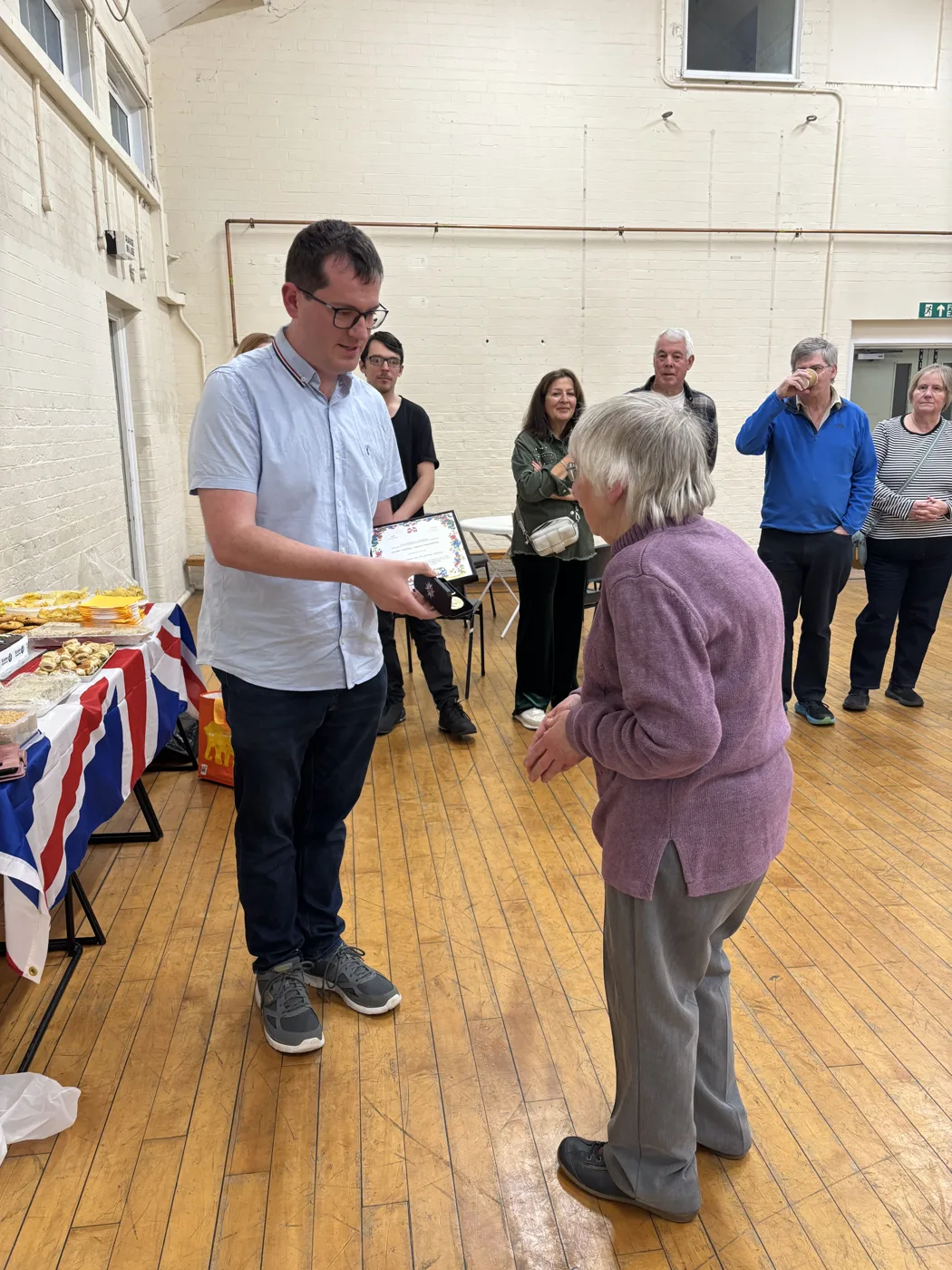 A man in a pale blue shirt holds a framed certificate and a small case as he makes a presentation to a woman in a lilac cardigan, in a hall with a wooden floor, while guests look on